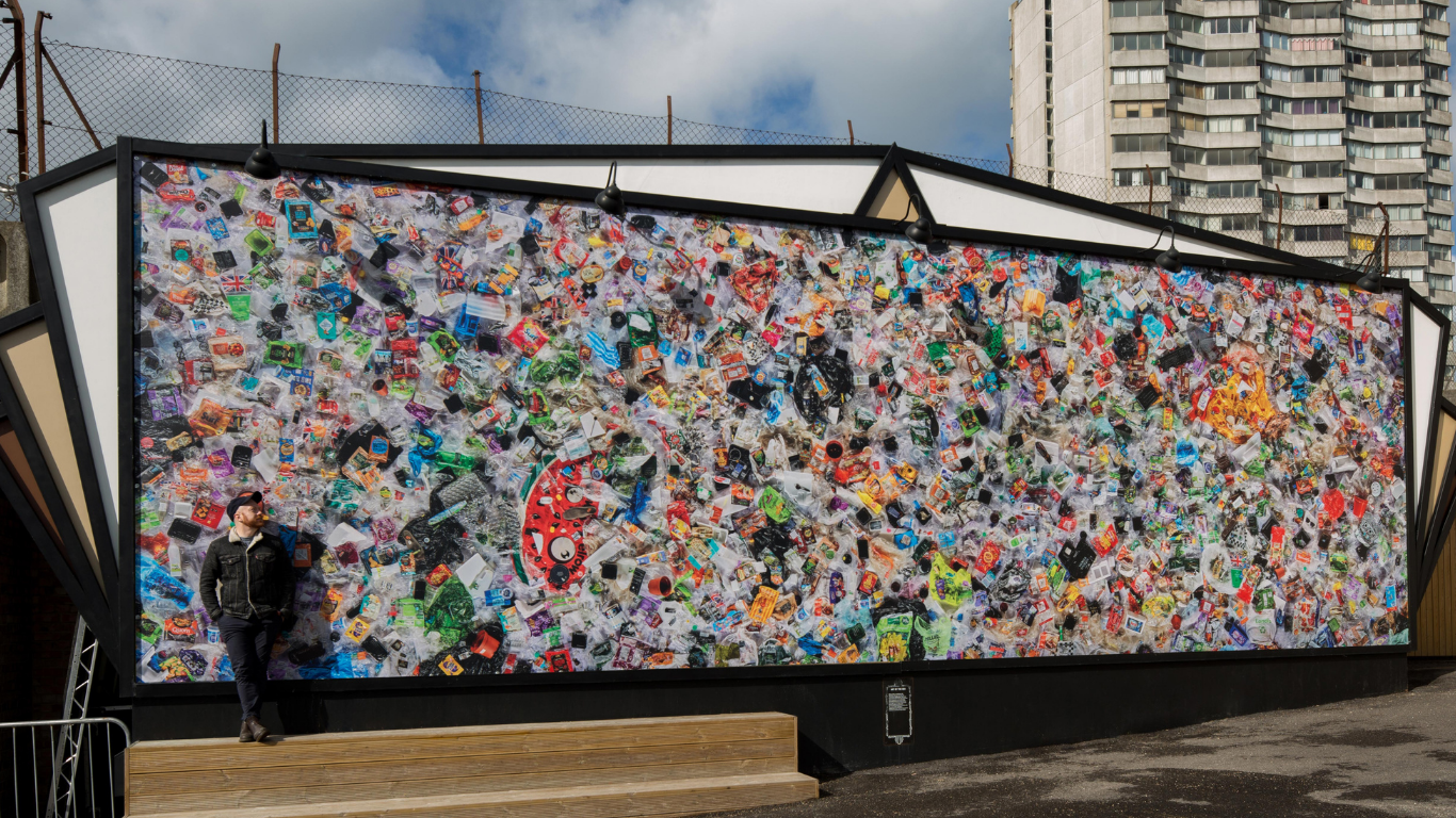 An artist stands in front of a billboard displaying thousands of pieces of plastic waste