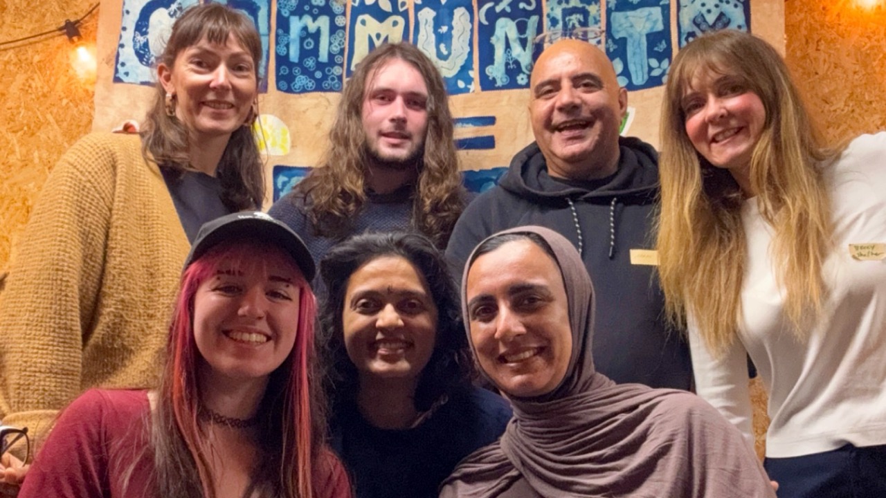Seven members of a community advisory group for The Big Plastic Count smile at the camera in front of a handmade sign reading "Community"